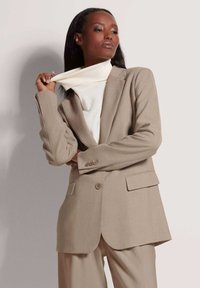Beige op maat gemaakte blazer met schouderflappen en twee frontzakken; voorzien van een enkele knopen sluiting en gecombineerd met een crèmekleurige coltrui.