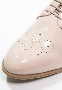 Chaussure en cuir verni rose clair avec un bout pointu, ornée de perforations décoratives en forme de cœur et une texture lisse et brillante.