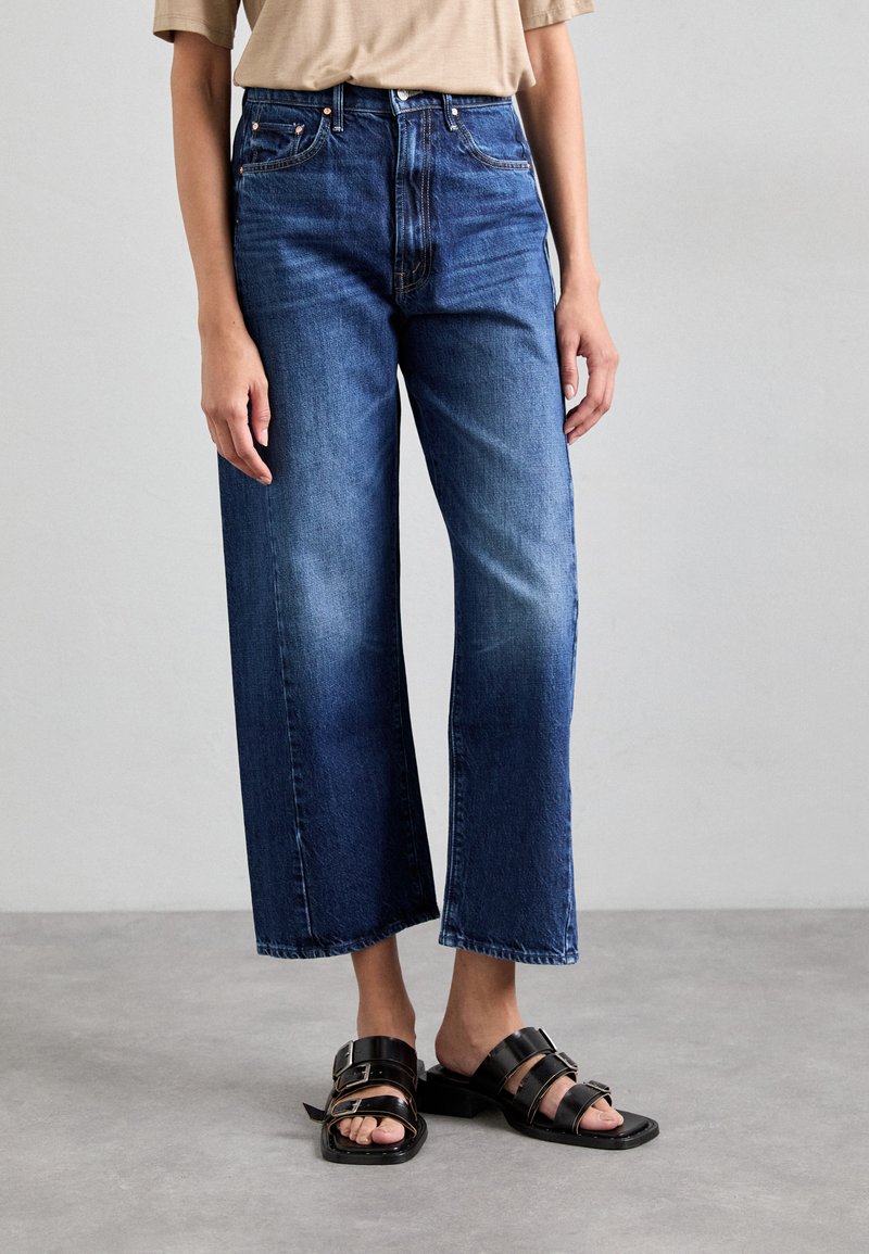Brede denim jeans in donkerblauw, met een hoge taille, vervaagde accenten, gestikte details en een schone zoom, gecombineerd met zwarte sandalen.