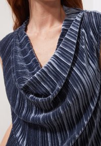 Námořnická modrá bezrukávová halenka vyrobená z texturovaného sametu, s drapovaným výstřihem a vertikálními sklady, s jemným lesklým povrchem.