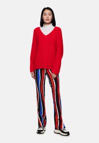 Roter, fuzzy Pullover mit V-Ausschnitt, kombiniert mit gestreiften Hosen in Rot, Blau, Schwarz und Weiß. Verfügt über eine gerippte Textur und ausgestellte Beine.