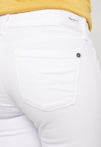 Jeans ajustados blancos con un bolsillo trasero visible, con costuras tonales y una etiqueta de marca en la cintura. Textura suave.