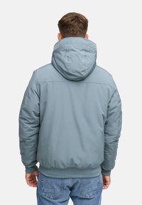 Man som bär en ljusblå vadderad jacka med huood och blå jeans, sedd bakifrån mot en enkel bakgrund.