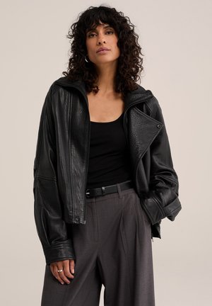 Femme aux cheveux bouclés foncés portant une veste en cuir noire, un débardeur noir, un pantalon gris et une ceinture noire, debout devant un fond uni.