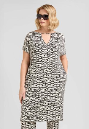 Donna con capelli biondi corti che indossa occhiali da sole neri e un vestito con motivo geometrico bianco e nero, maniche corte e tasche.