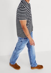 Koszulka polo w czarno-białe paski, niebieskie dżinsy oraz brązowe zamszowe kapcie. Mężczyzna idzie, prezentując ubrania codzienne i luźny krój.