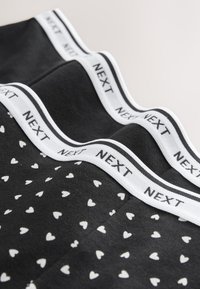 Sort undertøj med små hvide hjerter og hvide elastiske taljebånd med mærkenavnet "NEXT" i sort tekst.