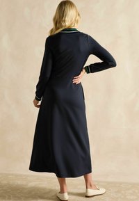 Marineblaues Langarmkleid mit Kragen, das grün-weiße Streifen aufweist, eine glatte Textur hat und eine ausgestellte Silhouette besitzt, kombiniert mit weißen Ballerinas.