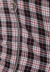 Rutigt tyg i svart, vitt och rött, med en synlig silverdragkedja och ringdragdetalj. Texturen verkar vävd och strukturerad.