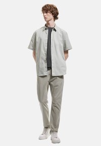 Camisa de botones de manga corta en verde claro sobre una camiseta gris oscuro, combinada con pantalones gris claro y zapatillas blancas. Diseño suave y casual.