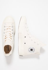 Hvite kanvas høy-hæl sneakers med svart logo-lapp, sølvfargede snørehull og lisser. Gummitåhette og teksturert såle.