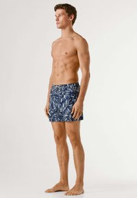 Ung mand, stående barfodet, iført blå badeshorts med hvide bladmotiver, vendt mod venstre mod en ensfarvet lys baggrund.