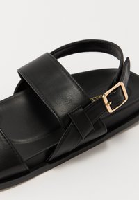 Sándalo de cuero negro con una cinta ancha y soporte en el talón, asegurado con una hebilla en tono dorado. Textura suave y plantilla acolchada.