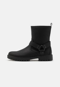 Botas negras de tobillo con parte superior de cuero texturizado, que cuentan con una correa lateral con adornos metálicos y una suela de goma con tacos para mayor tracción.