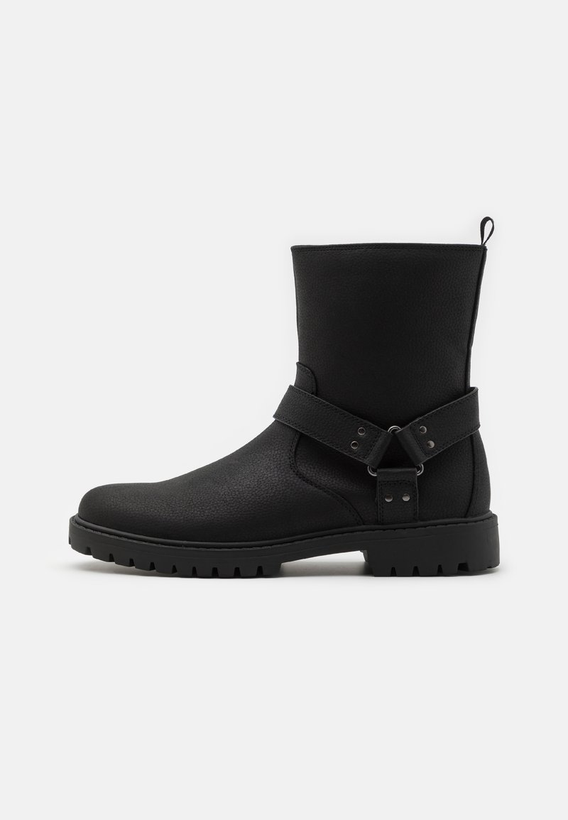 Botas negras de tobillo con parte superior de cuero texturizado, que cuentan con una correa lateral con adornos metálicos y una suela de goma con tacos para mayor tracción.