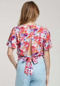 Blouse florale dans des teintes vives d'orange, de rose et de violet. Tissu léger avec des manches courtes et un détail de nœud dans le dos. Dos ouvert avec fermeture à bouton.