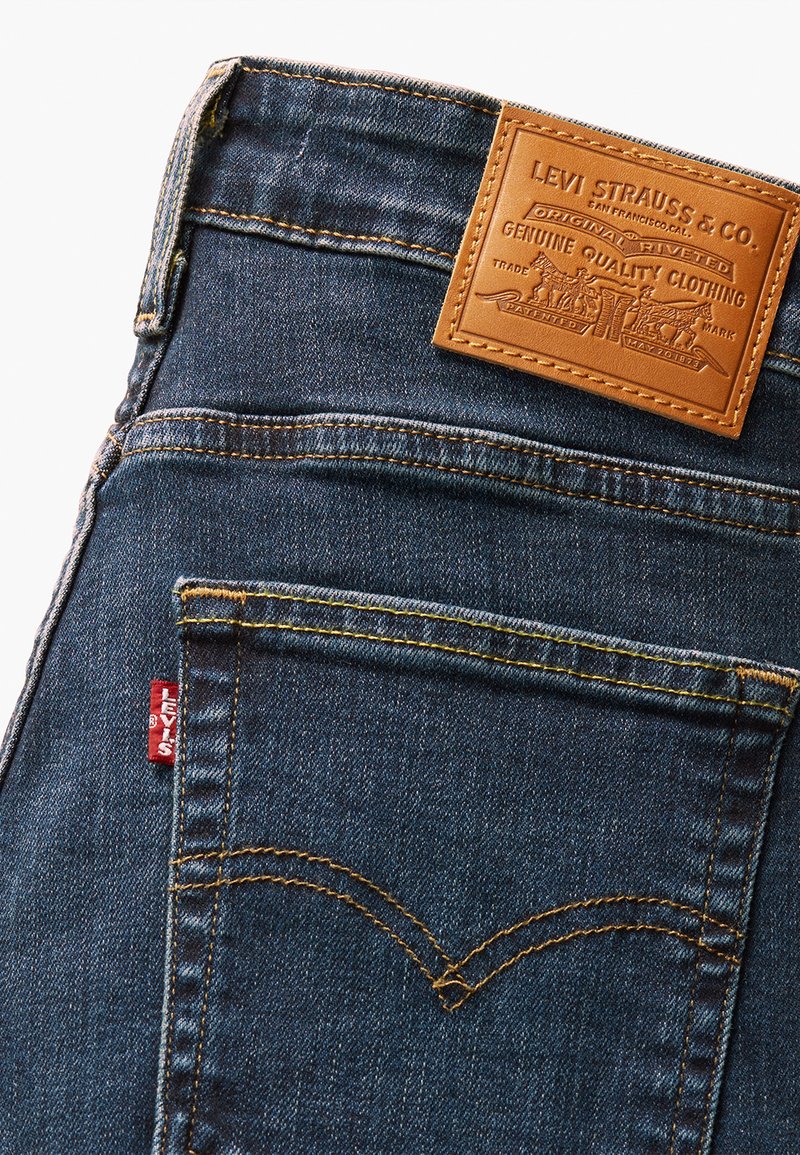 Vaqueros de mezclilla en azul oscuro con costuras amarillas, con una etiqueta de cuero marrón en la cintura y una etiqueta roja en el bolsillo trasero.