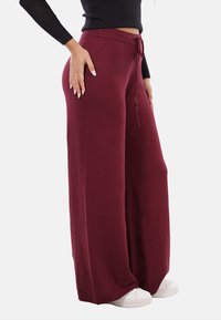 Pantaloni della tuta a gamba larga di color bordeaux realizzati in tessuto morbido; presentano una vita con coulisse e sono abbinati a sneakers bianche, mostrando un fit rilassato.