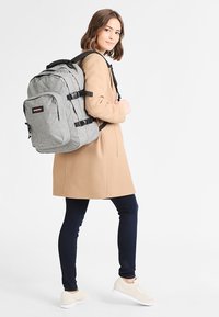 Mochila de tela gris con correas ajustables, múltiples compartimentos y acentos en negro. El modelo lleva un abrigo beige y jeans oscuros.