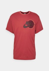 Punainen Nike-paita, jossa musta swoosh-logo ja pyöreä keltainen teksti vasemmassa rinnassa, lyhyet hihat ja pyöreä kaulus.