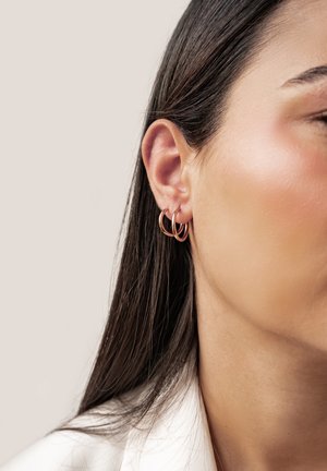 Nahaufnahme eines rechten Ohrs einer Frau mit zwei kleinen, dünnen Rosengold-Creolen und glattem dunkelbraunem Haar vor hellem Hintergrund.