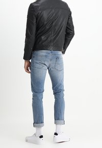 Man in zwarte leren jas, lichtblauwe opgerolde jeans, witte sokken en witte sneakers, staand met de rug naar de camera tegen een witte achtergrond.