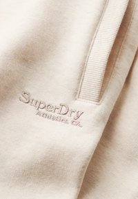 Tissu beige au toucher doux, avec un logo brodé "SuperDry Athletics, CA" en marron clair. Accents côtelés visibles en haut.