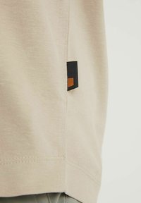 Tissu beige avec une petite étiquette rectangulaire noire et orange cousue près de la couture, à côté d’un ourlet doublement piqué.