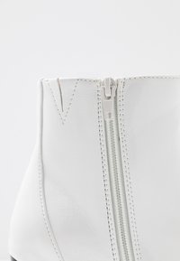Weißer Knöchelstiefel mit einem zentralen Reißverschluss, glattem synthetischem Material, verstärkten Nähten und einem runden Zehendesign mit einem leichten Glanz.