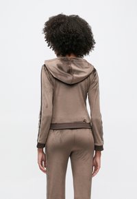 Survêtement en velours marron avec capuche, présentant des bandes blanches sur les côtés. Texture lisse, coupe ajustée avec poignets et taille côtelés.