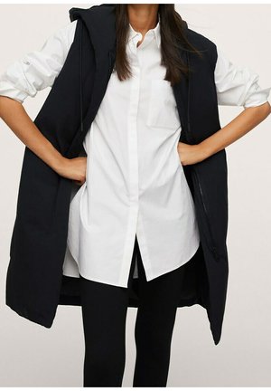 Vrouw draagt een lange zwarte mouwloze hoodie-vest over een wit overhemd met opgerolde mouwen en zwarte broek.