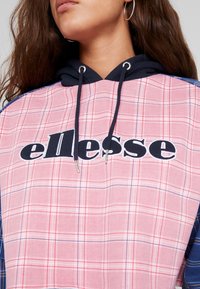 Felpa con cappuccio a quadri rossi e rosa con dettagli blu navy e occhielli argentati. Presenta un logo nero "ellesse" ben visibile sul davanti. Tessuto texture.