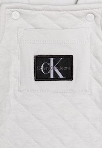Biały pikowany materiał z czarną prostokątną naszywką z napisem "Calvin Klein Jeans" oraz stylizowanym logo "CK", ozdobiony dwoma zatrzaskami.