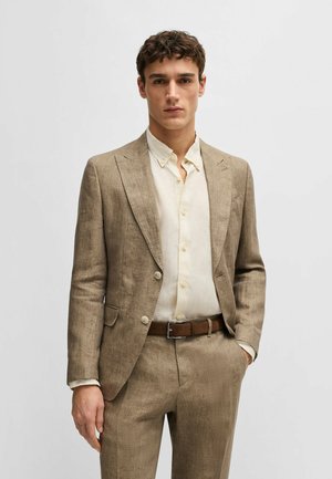 Costume en lin marron clair avec une coupe ajustée, blazer à simple boutonnage et pantalon à plis plats, associé à une chemise beige à boutons et une ceinture marron.
