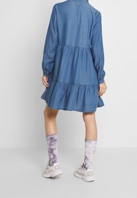 Denimmekko pitkillä hihoilla ja kerroksellisella röyhelöhelalla. Yhdistetty purppuranvärisiin taitaustossuihin ja valkoisiin lenkkareihin. Napitettu hihansuu, rento malli.