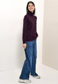 Maglione dolcevita color bordeaux abbinato a jeans larghi blu e sneakers grigie, su uno sfondo neutro che mette in risalto i colori dell'outfit.