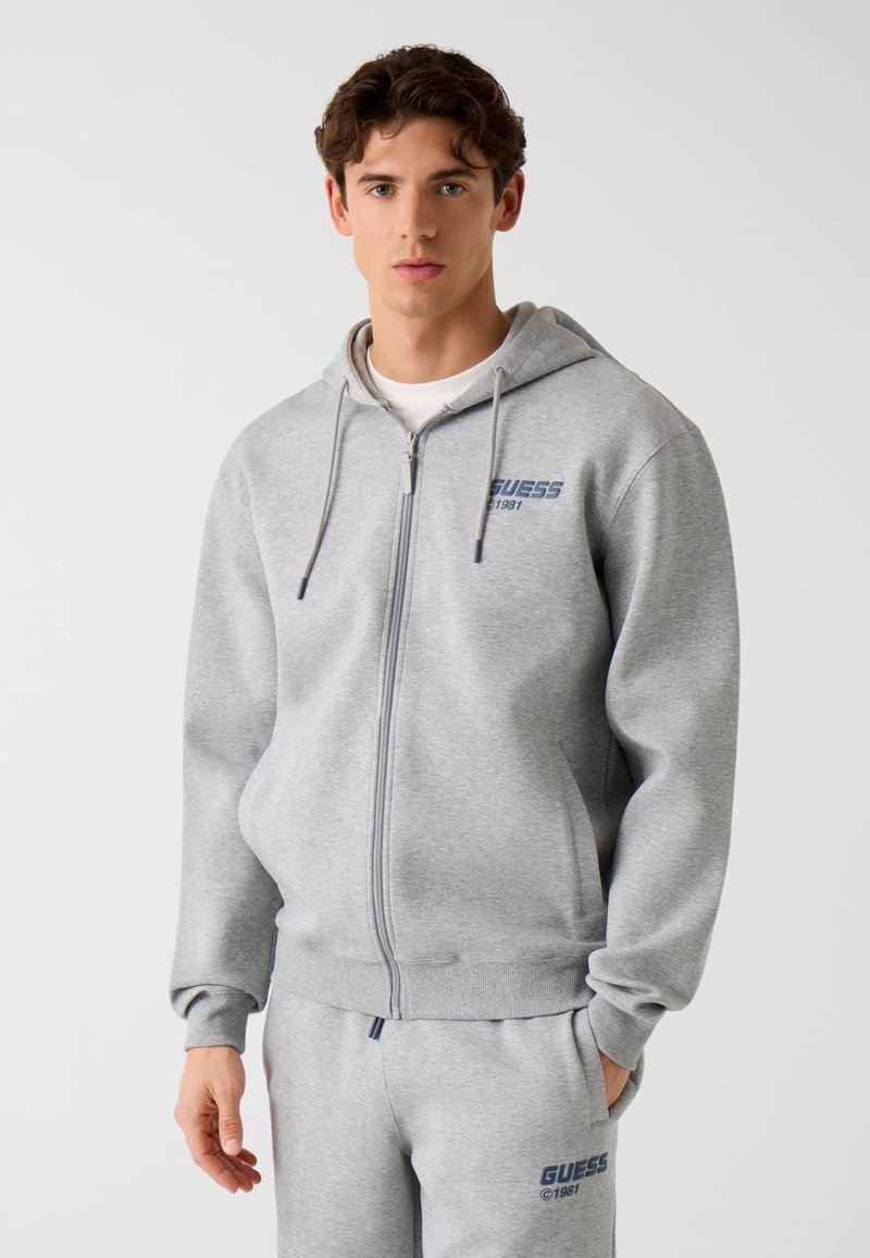 Sweat à capuche gris zippé en tissu doux, avec une capuche à cordon de serrage et un logo bleu marine sur la poitrine. Texture lisse, coupe ajustée.