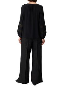 Schwarze Bluse mit langen Ärmeln und Spitzen-Einsätzen; kombiniert mit lockeren schwarzen Hosen. Glatte Textur, lässige Passform.
