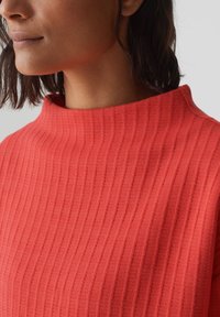 Pull en maille côtelée de couleur corail vif, avec un col montant et un tissu texturé. Gros plan mettant en avant le motif de point et la couleur unie.
