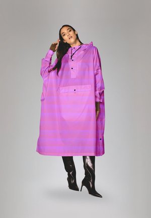 Vrouw draagt een loszittende felroze regenponcho met zwarte knopen en zwarte hoge hakken tegen een effen achtergrond.