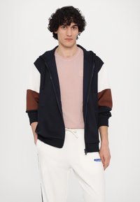 Jūrinio stiliaus zip-up džemperis su baltais ir ruda akcentais, dėvimas virš šviesiai rožinės spalvos marškinėlių, derinamas su baltais kelnėmis. Minkšta medžiaga, kasdieniškas dizainas.