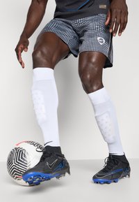 Schwarze und blaue Fußballschuhe, graue gemusterte Shorts, weiße Schienbeinschoner und ein schwarz-weißer Fußball vor einem hellen Hintergrund.