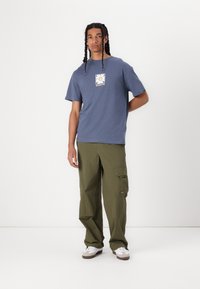 Tricou din bumbac albastru cu mâneci scurte și un imprimeu grafic, asortat cu pantaloni cargo verzi măslinii, care au buzunare și o croială relaxată.