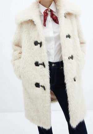 Kobieta ubrana w biały, puszysty płaszcz z czarnymi guzikami na zatrzask, białą koszulę zapinaną na guziki, czerwoną chustę w kwiaty na szyi oraz ciemne dżinsy.