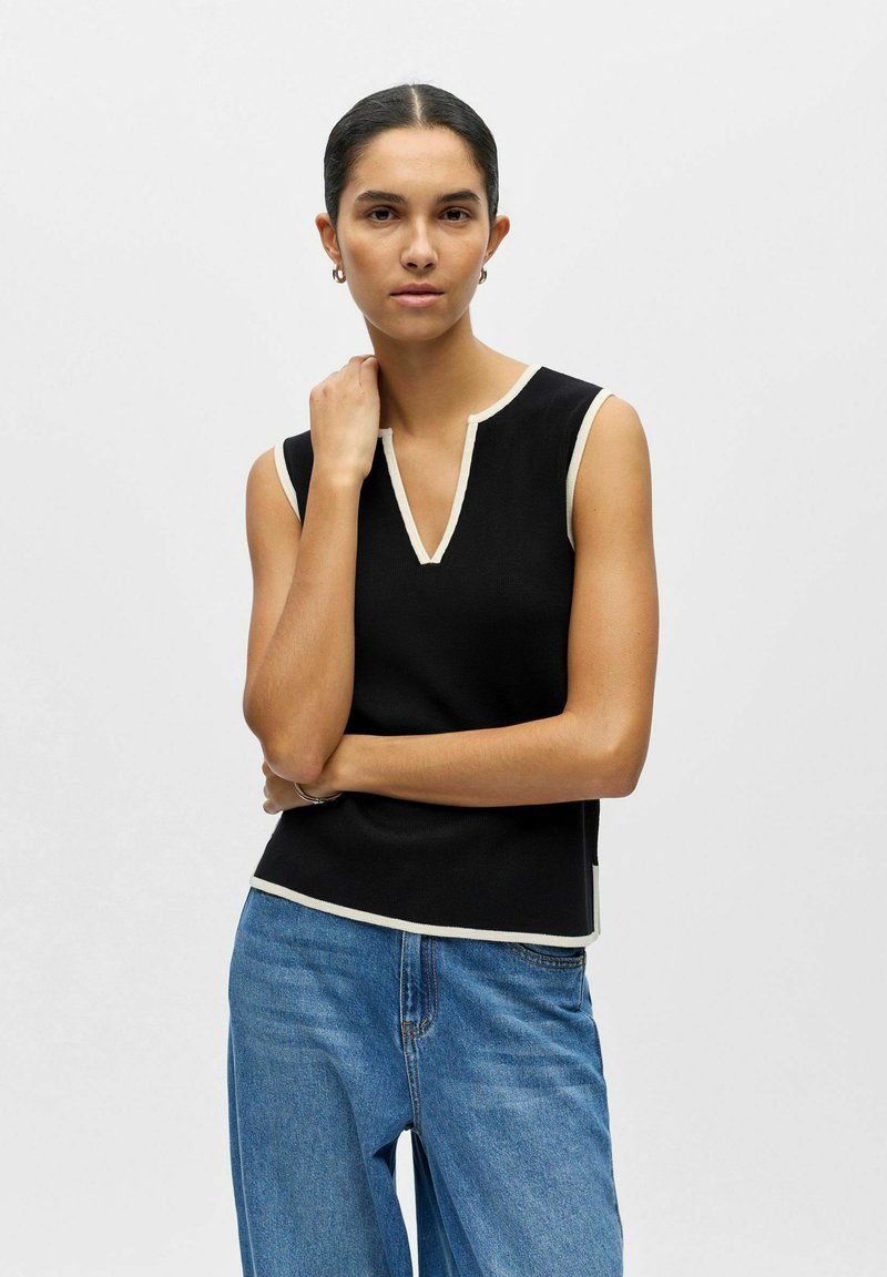 Femme aux cheveux foncés attachés en arrière, portant un haut noir sans manches avec un liseré blanc et un jean bleu, posant avec un bras croisé et la main sur le cou.