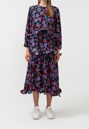 Frau trägt ein schwarzes Midikleid mit Blumenmuster in Pink, Türkis und Rot, mit Rüschenlagen und weißen Turnschuhen, steht vor einem weißen Hintergrund.