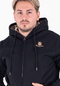 Felpa nera con zip, caratterizzata da una fodera interna a scacchi, cordini regolabili e un logo sul petto. Tessuto morbido.