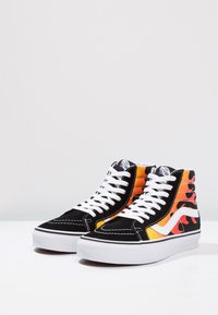 Højtop sneakers med sort ruskind, orange og gul flamme mønster, hvide snørebånd og hvid gummisål. Side logo lavet af hvidt materiale.