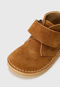 Zapato de ante marrón con punta redondeada, lados abiertos y una correa de velcro. Presenta costuras blancas en contraste y una suela de goma para mayor tracción.