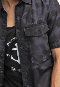 Mężczyzna ubrany w czarną, krótko rękawową koszulę w kamuflaż, rozpiętą na czarny top z białym wzorem kotwicy i napisem.